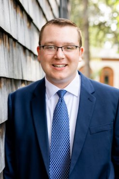 Garrett Platt in blue suit, white shirt, blue tie and glasses, stands beside a house and smiles.